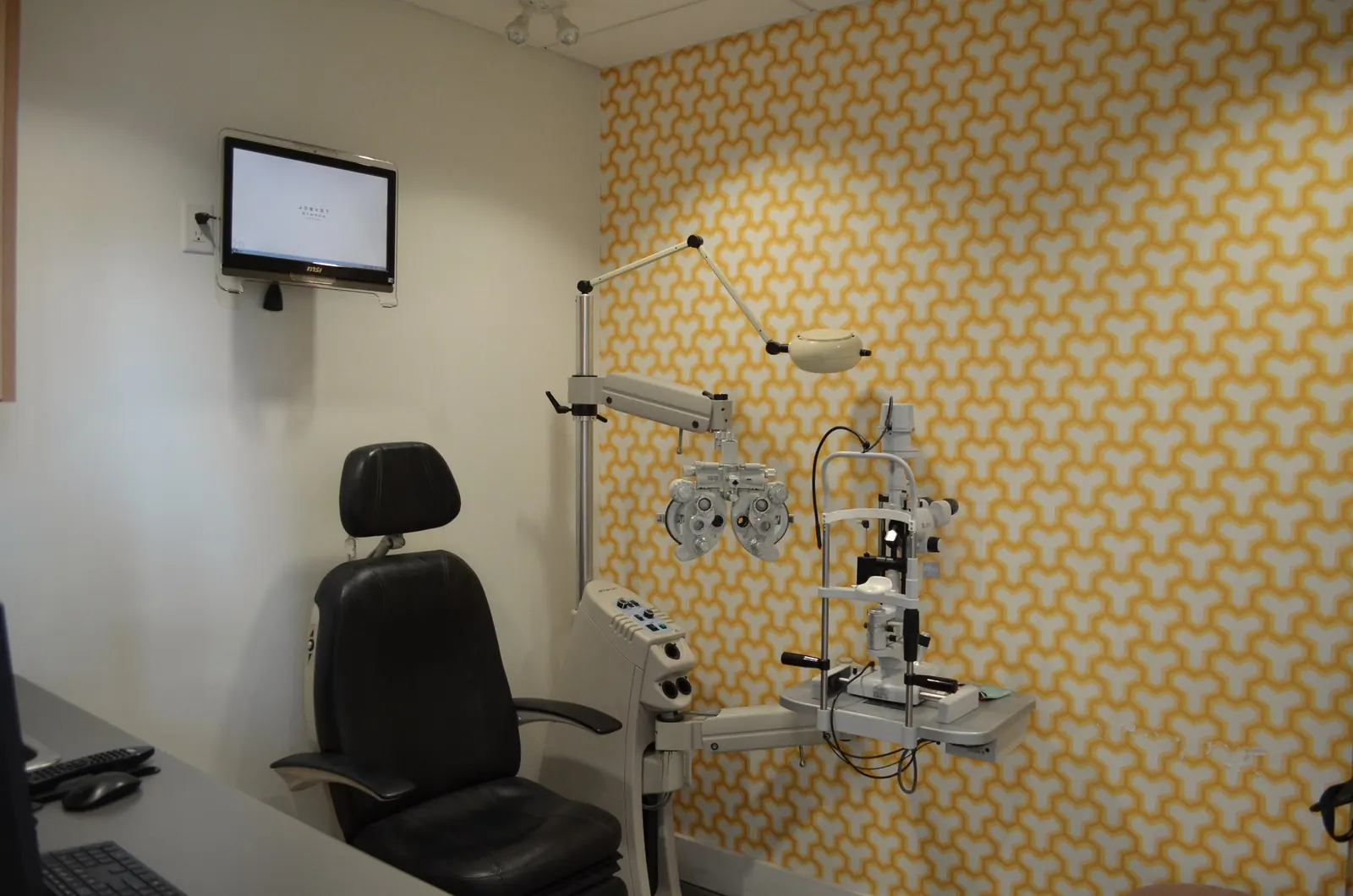 An exam room at The EyeSite: exam chair, phoropter and slit lamp in front of patterned wallpaper